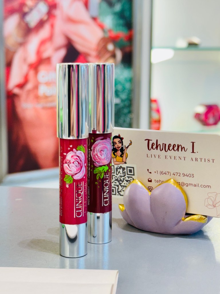 Two Clinique lip products with floral designs placed on a gray surface, beside a business card for live event artist Tehreem I, along with a decorative purple lotus-shaped holder.