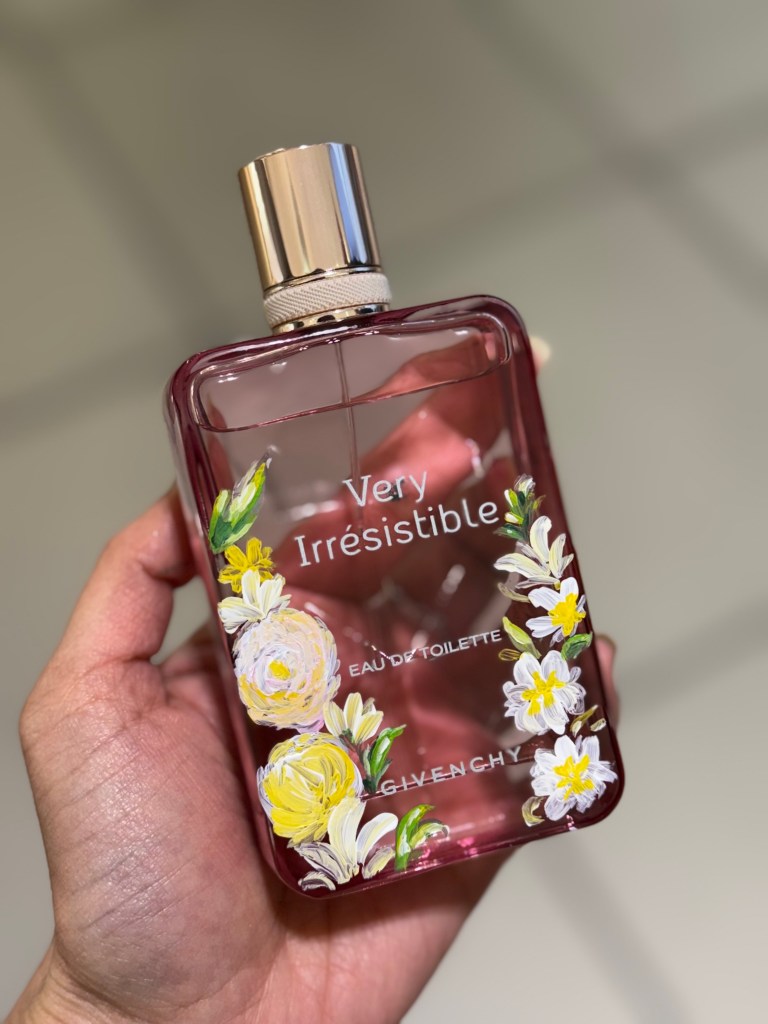 A person's hand holding a pink perfume bottle with floral decorations, labeled 'Very Irresistible' by Givenchy.